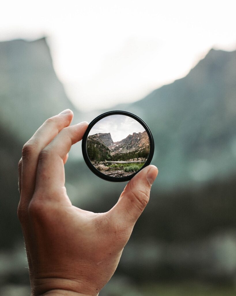 a white hand holds up a small round mirror that shows the clear refleciton of the mountains, the background is blurred