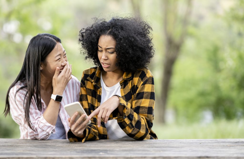 Cheerful Black and Asian lesbian women sitting at table using smartphone outdoors